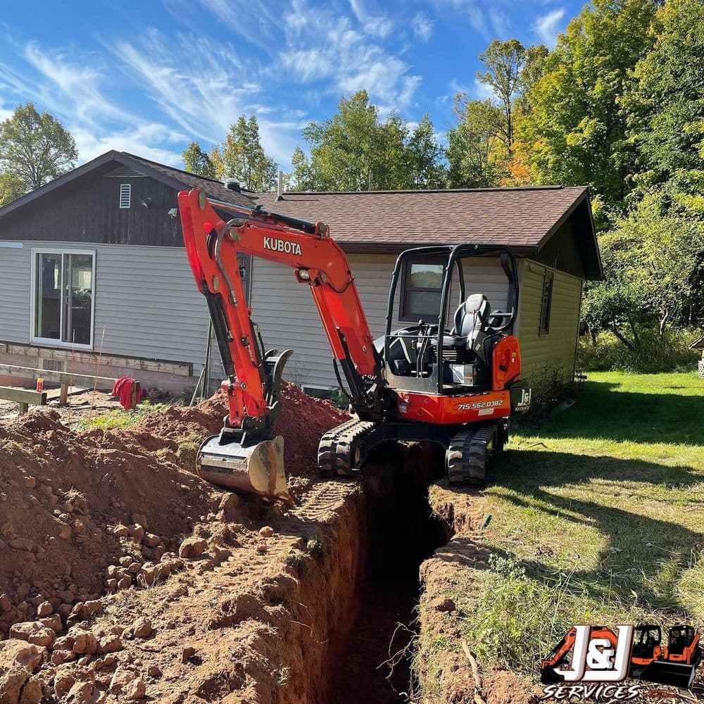 Kubota excavator digging a trench beside a house on a sunny day. J&J Services logo visible.