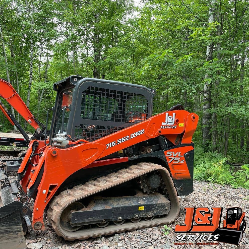 Orange compact track loader by J&J Services in a wooded outdoor setting.