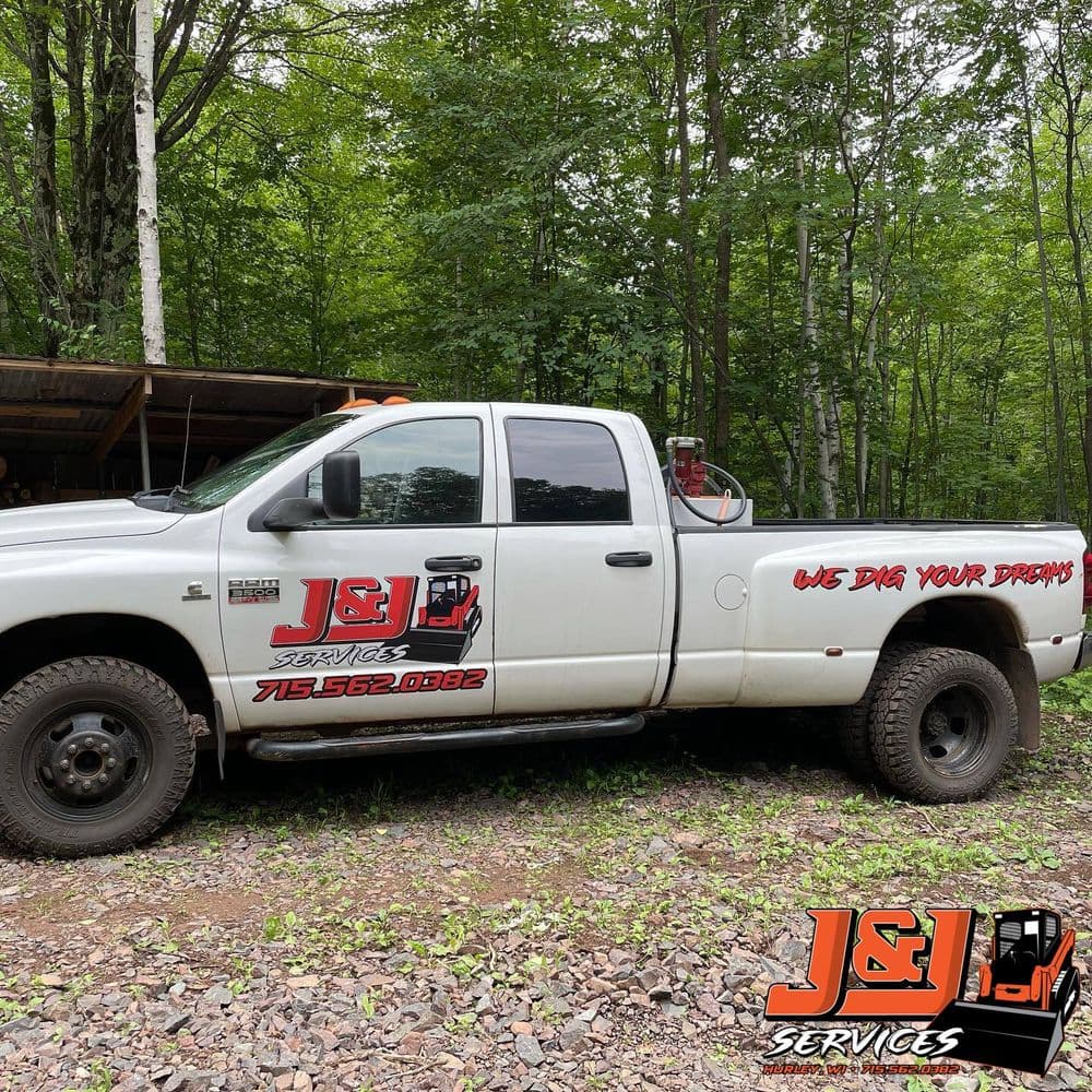 White pickup truck featuring J&J Services logo and "We Dig Your Dreams" slogan in a wooded area.