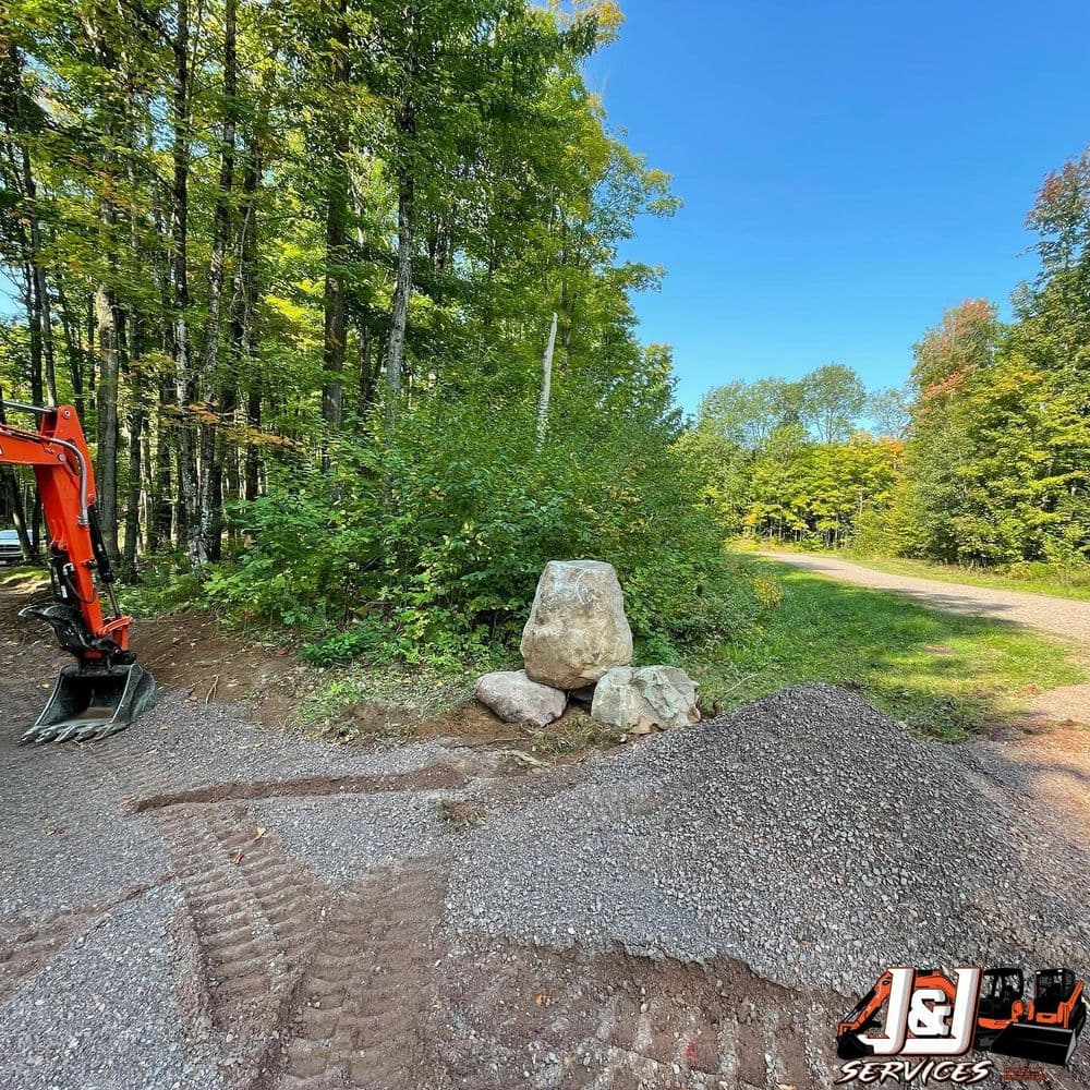 Excavator and gravel beside large rock in wooded area during landscaping project.