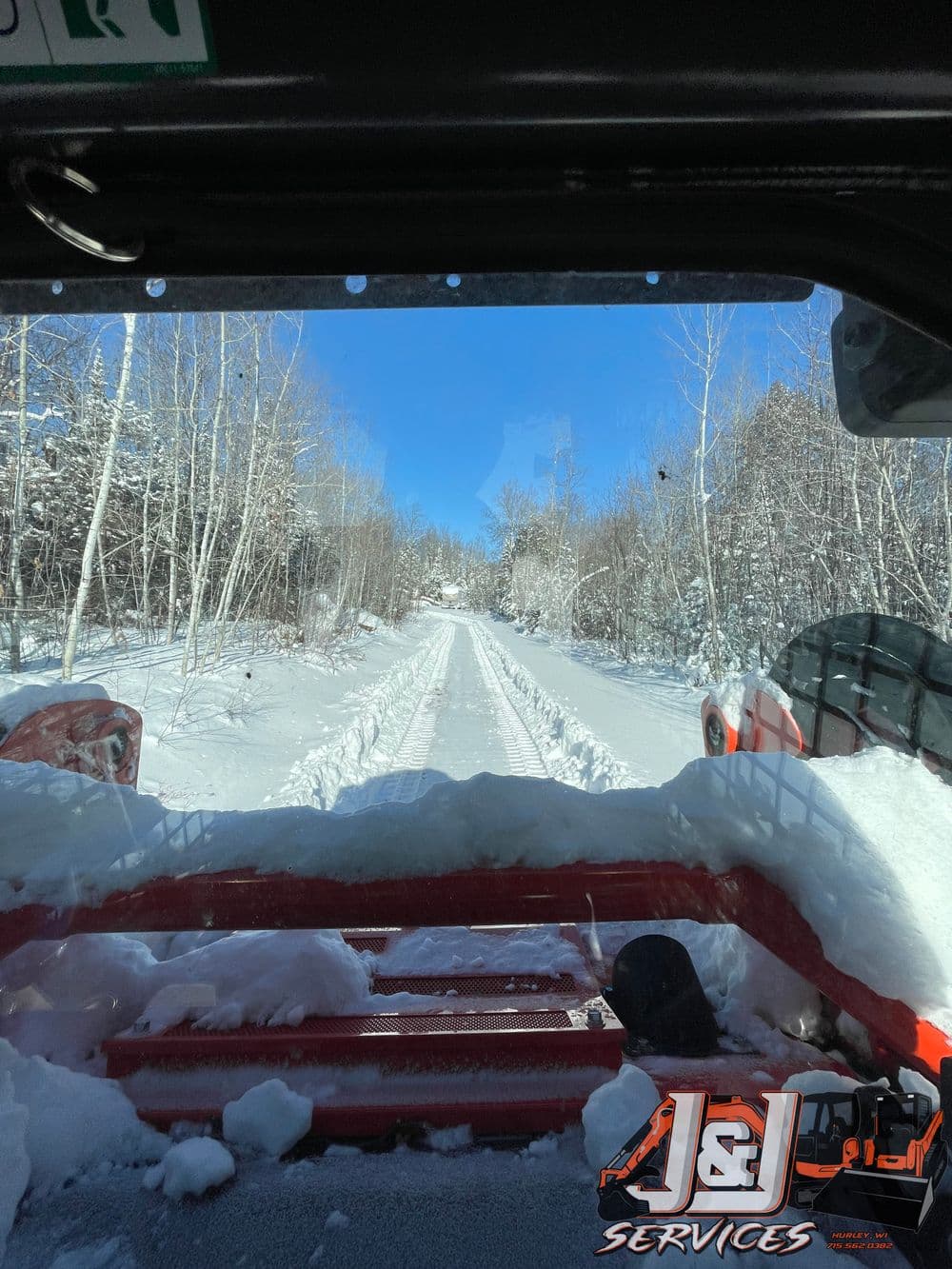 View from a snowplow clearing a snow-covered road lined with trees under a blue sky.