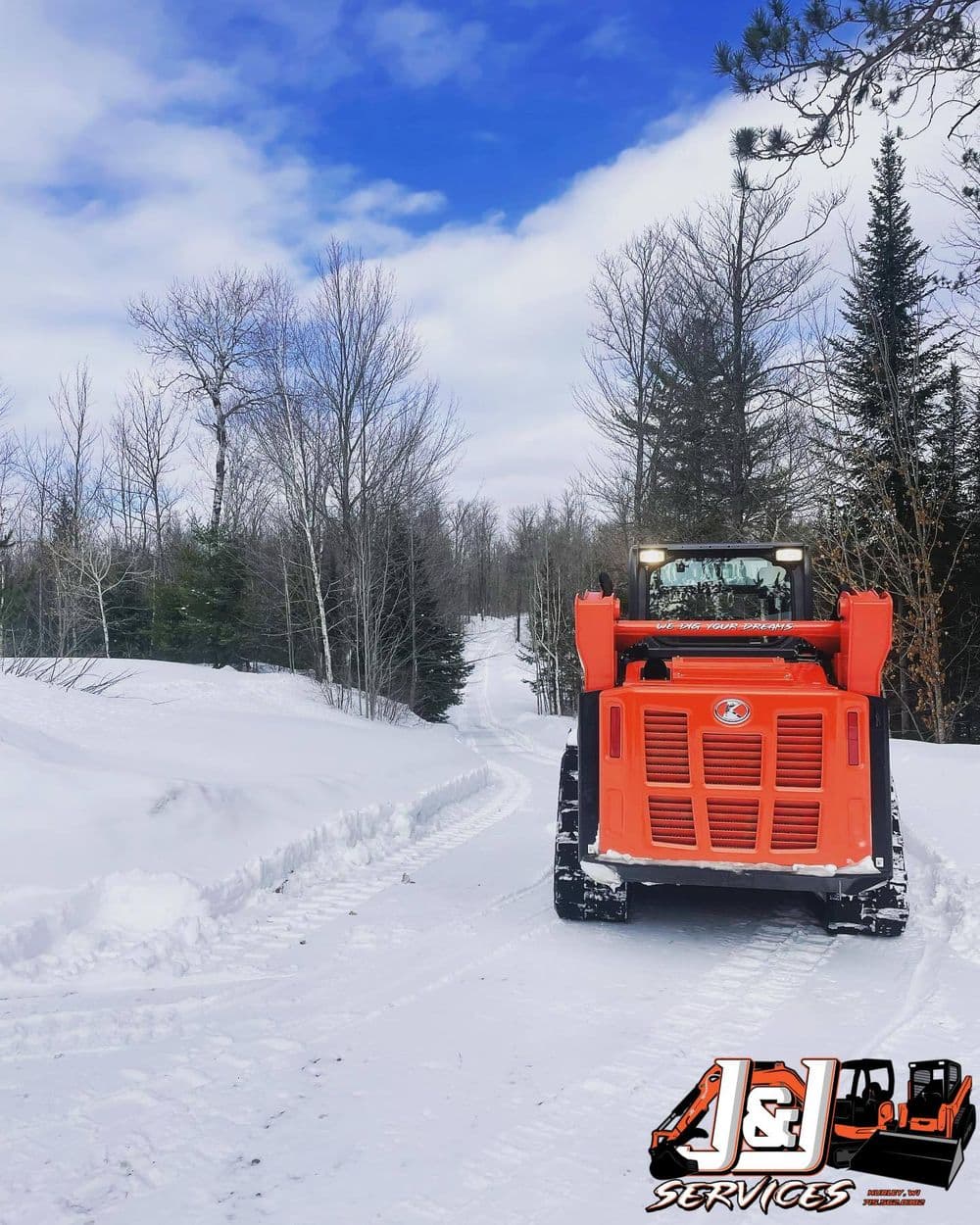 Orange snow plow clearing a snowy path through a forested area on a bright winter day.