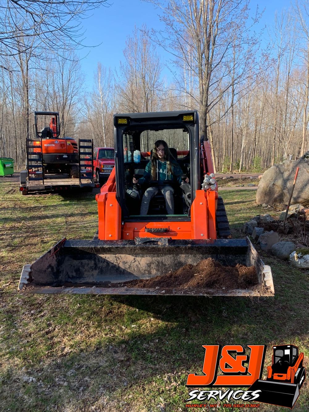 Person operating a tractor with a front loader in a wooded area, J&L Services logo visible.