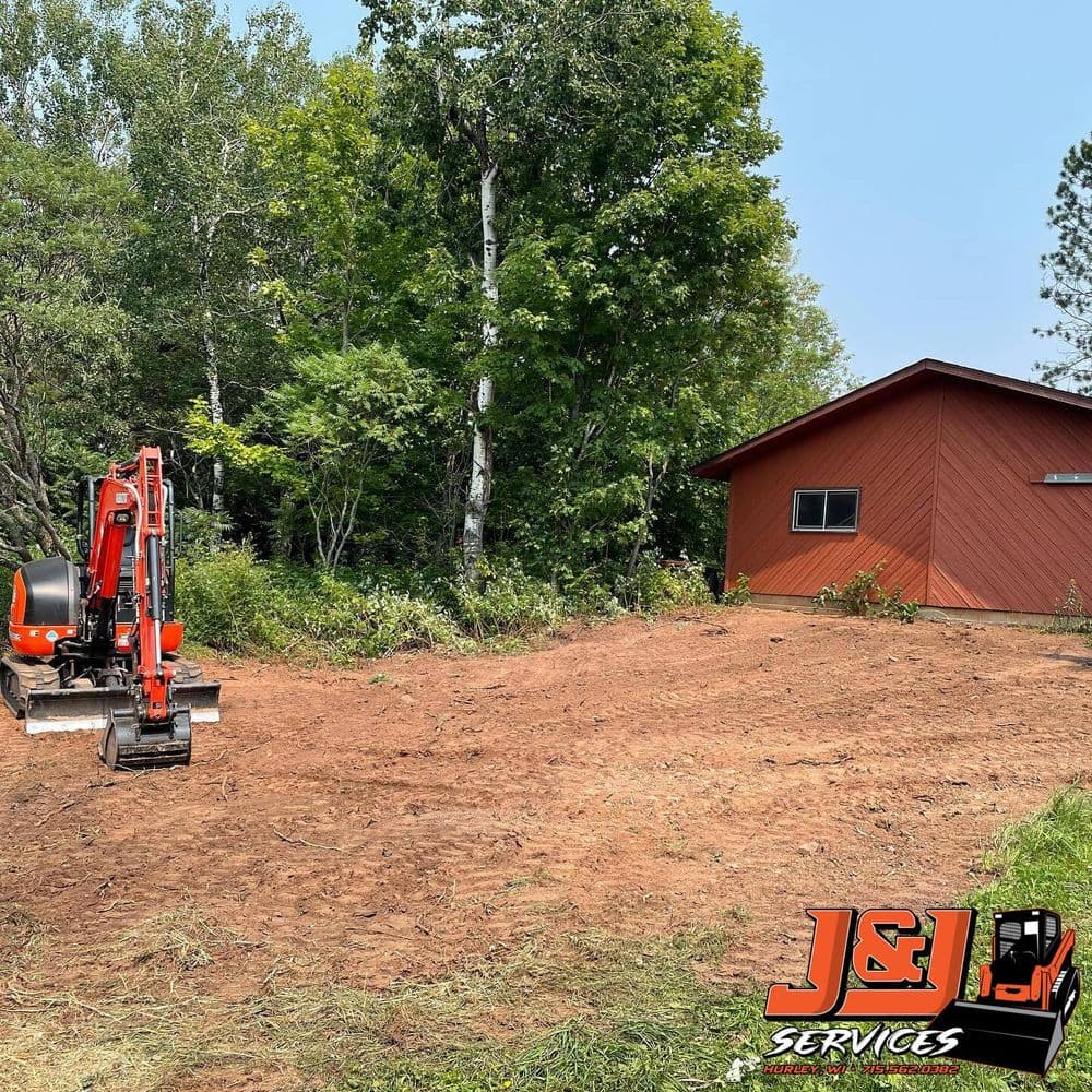 Excavator on cleared land beside a red service building in a wooded area. J&L Services logo.