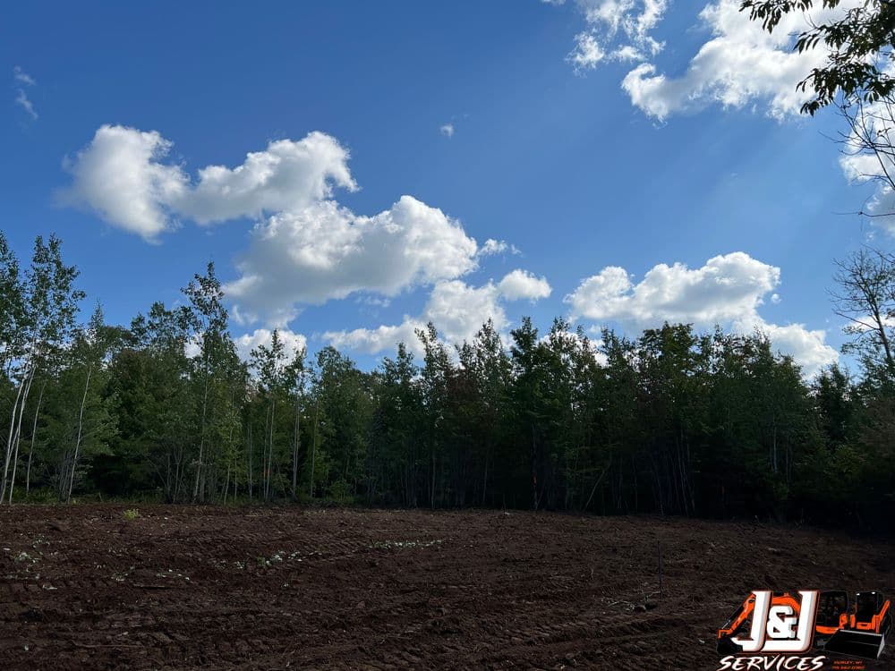 Cleared land with a blue sky and white clouds, surrounded by green trees. J&L Services logo.