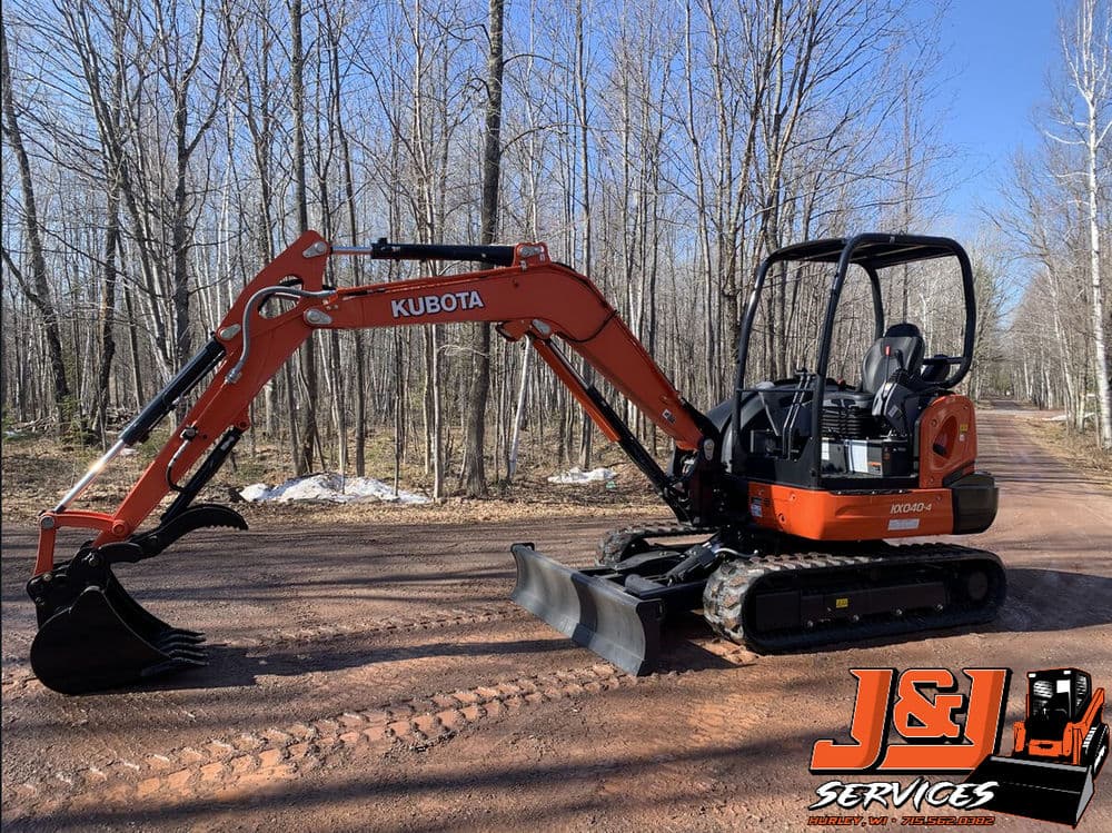 Kubota excavator on a dirt road in a forested area, branded with J&L Services logo.