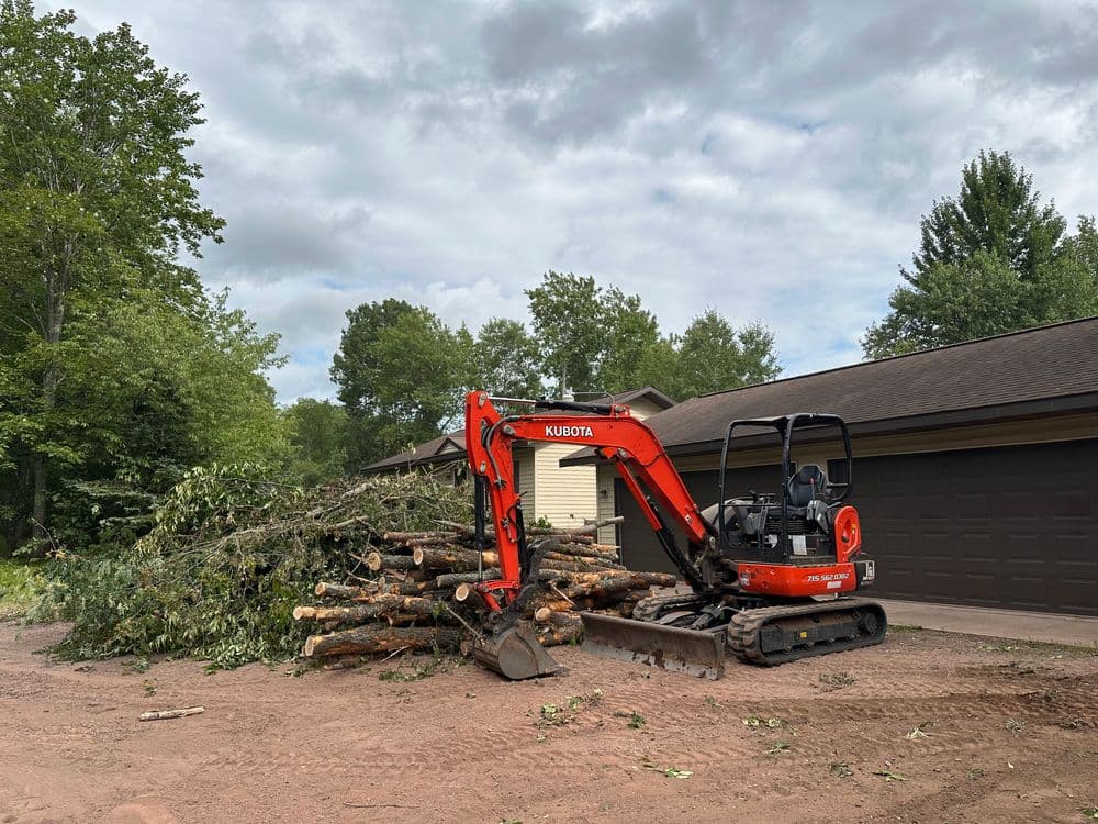 Kubota mini excavator near a pile of cut tree logs outside a house in a wooded area.