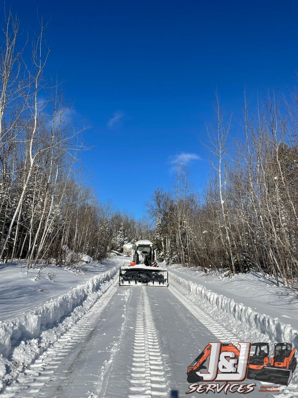 Snow plow clearing a snowy road with blue sky and trees in the background. J&L Services logo.