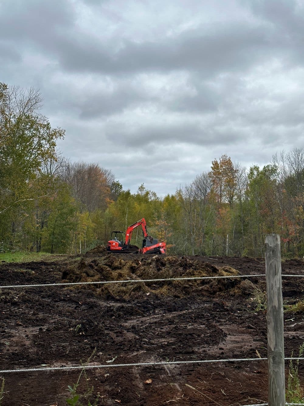 Excavator working on a muddy field with autumn trees and overcast sky in the background.