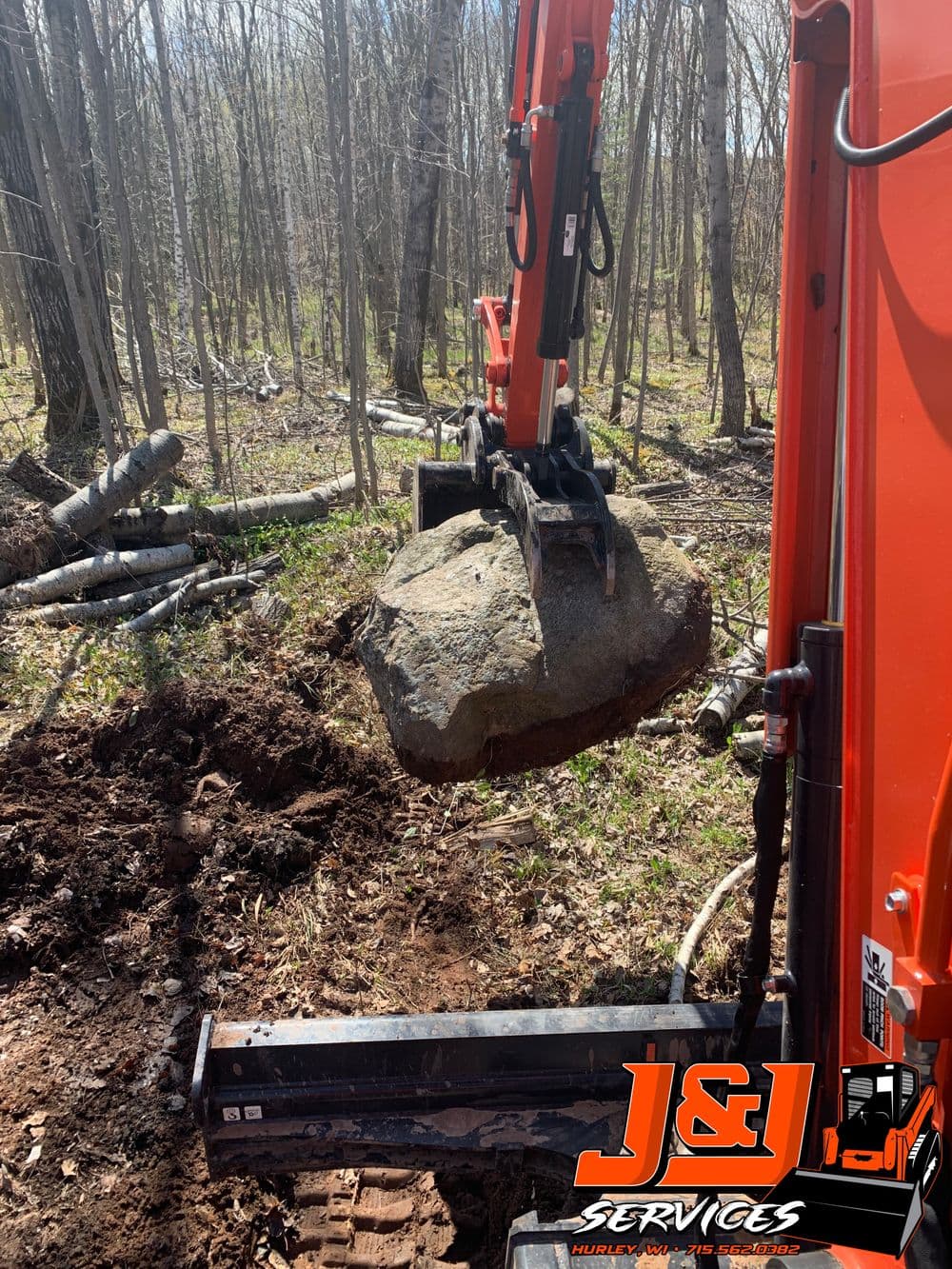 Excavator lifting a large boulder in a forested area, J&L Services logo visible.