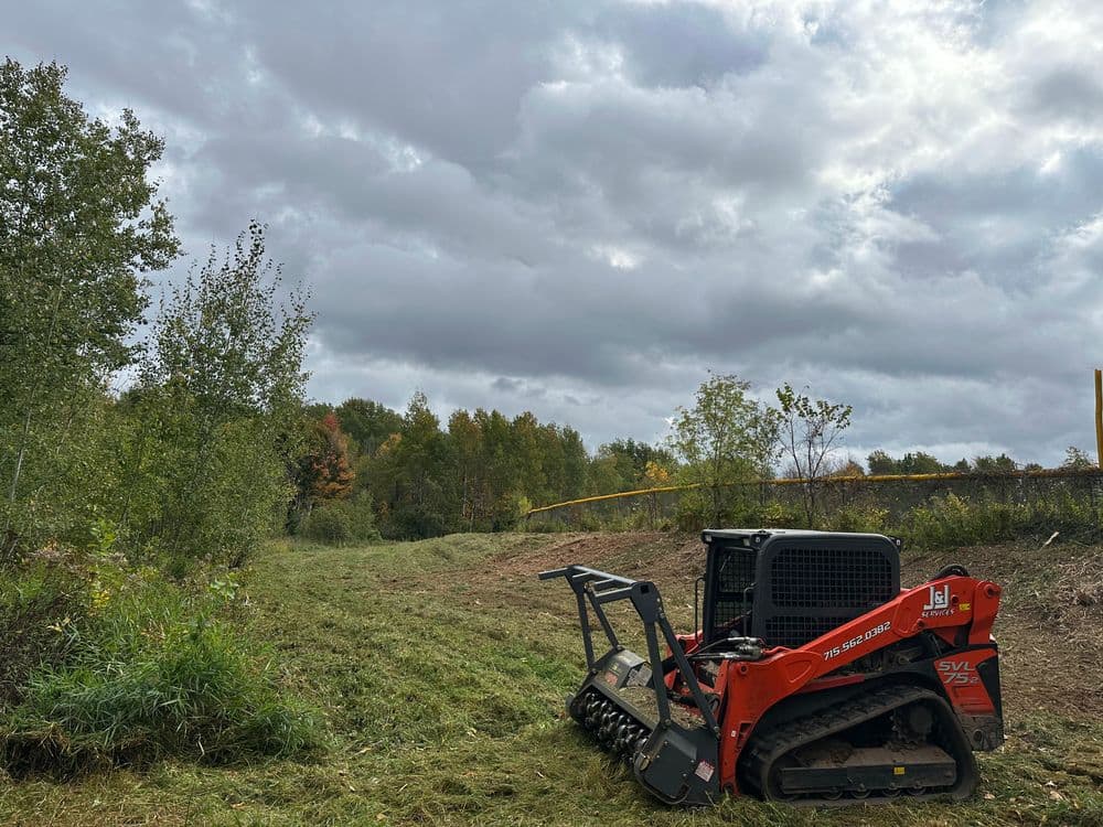Track loader parked on a cleared landscape under a cloudy sky, surrounded by trees.