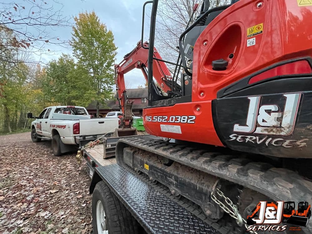 Excavator on trailer near truck in autumn landscape, J&L Services branding visible.