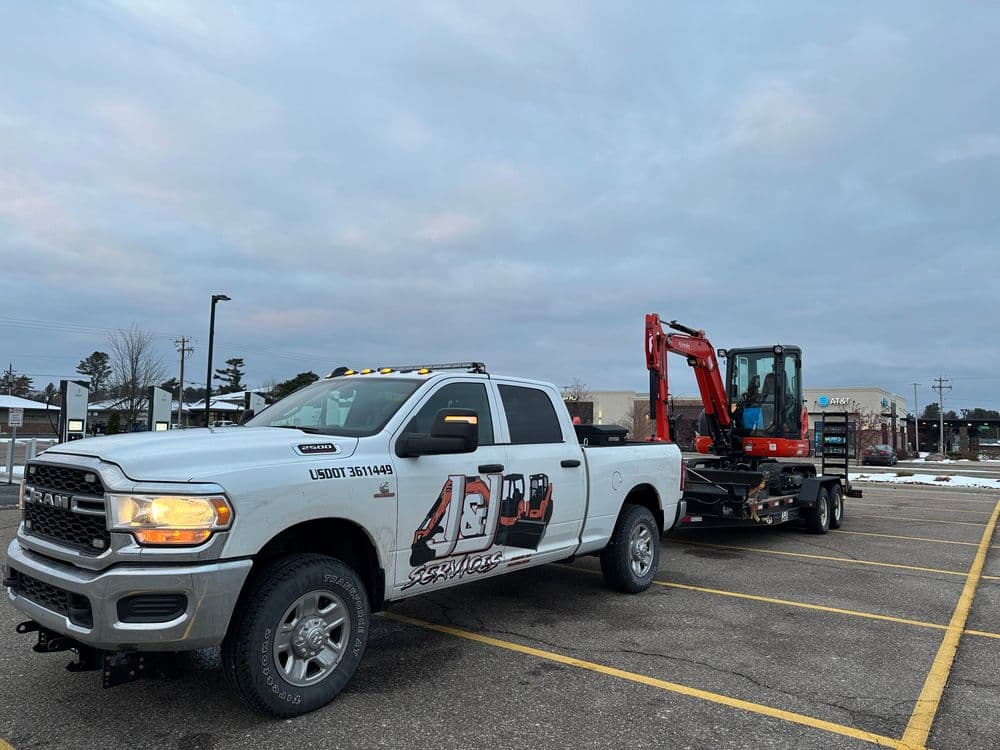 White pickup truck with logo towing a mini excavator on a trailer in a parking lot.