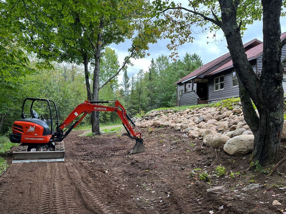 Excavator working on landscaping near a cabin surrounded by trees and rocks.