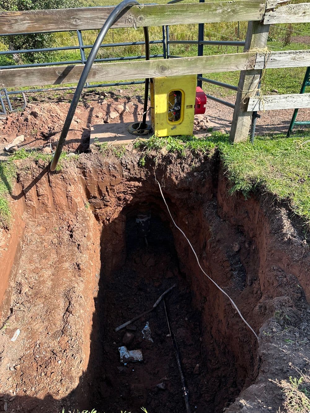 Excavation site showing a deep trench near a wooden fence and utility equipment.