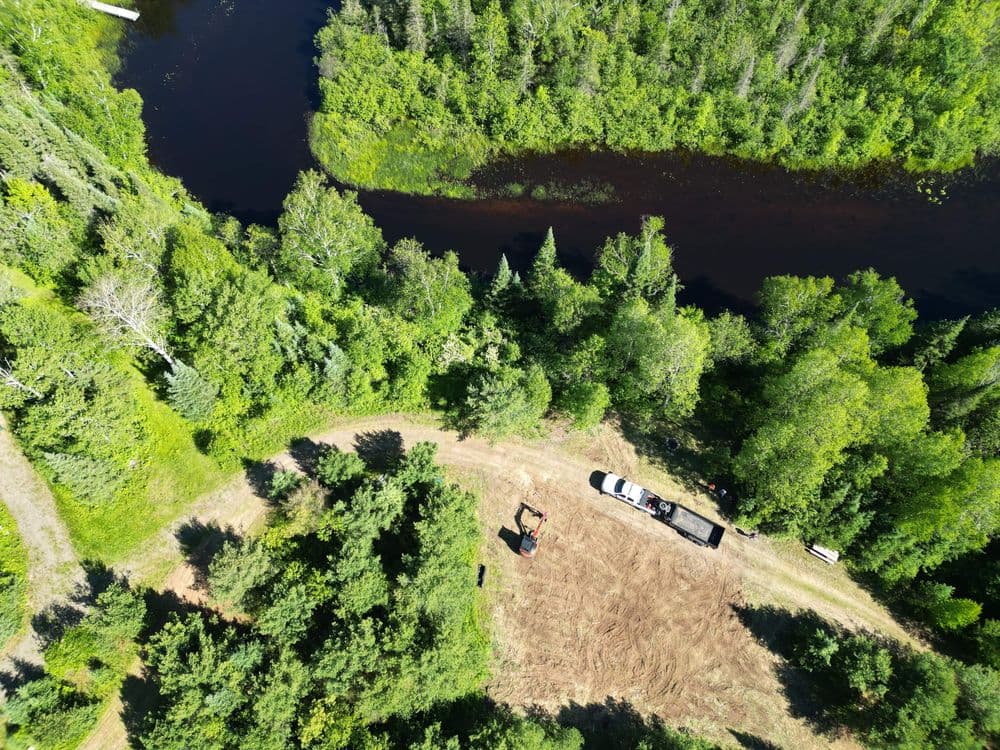 Aerial view of a forested area with a winding river and vehicles on a dirt path.