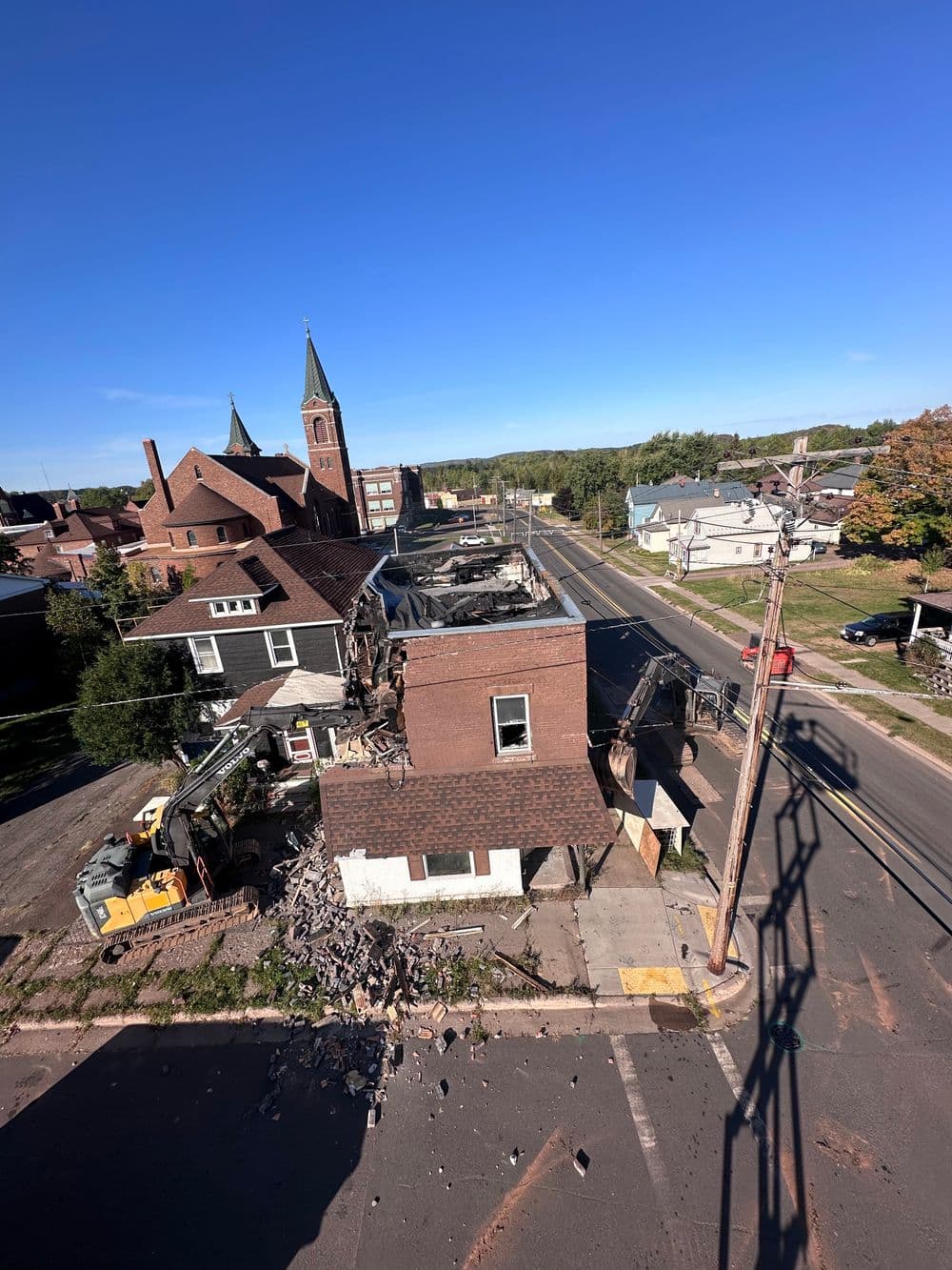 Demolished building with heavy machinery and church steeple in background, clear blue sky.
