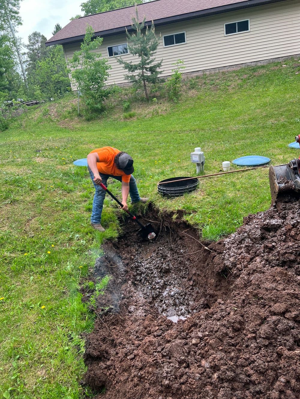 Person digging in the ground with a shovel in a garden, near a house and green trees.
