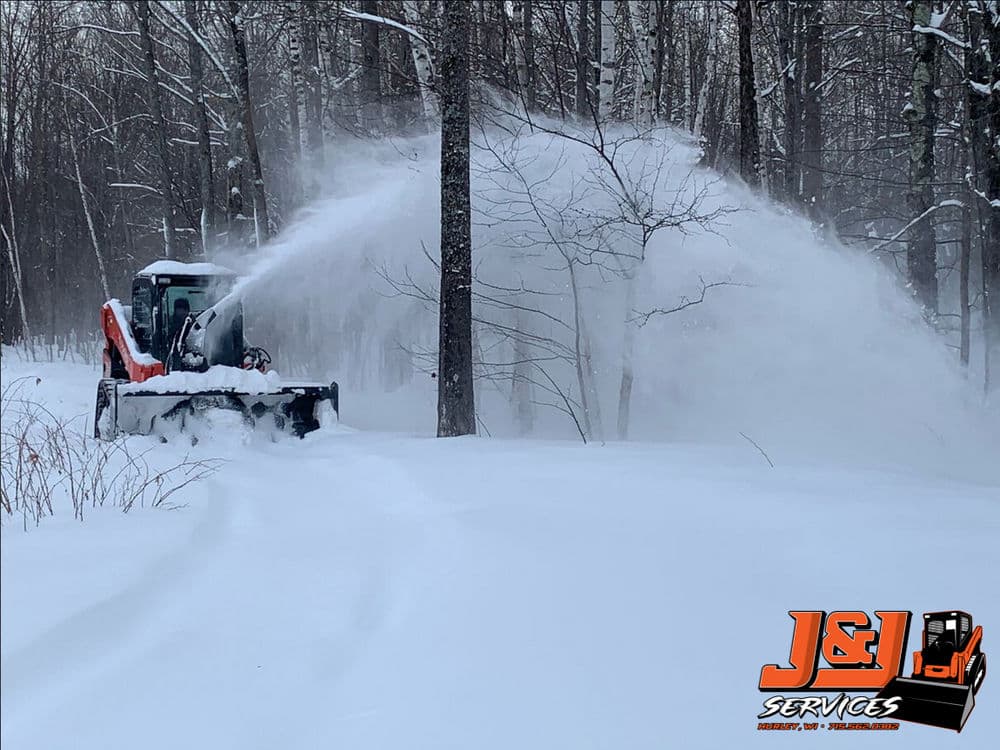 Snow removal equipment clearing a snow-covered forest path in winter. J&L Services logo visible.