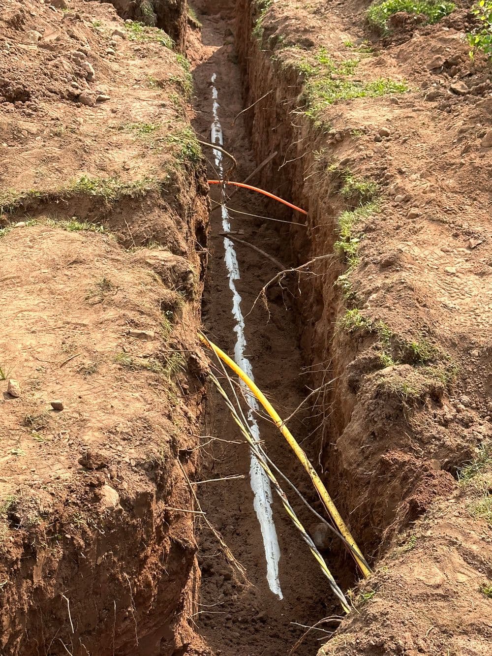 Excavated trench showing orange and yellow utility lines on unpaved ground.