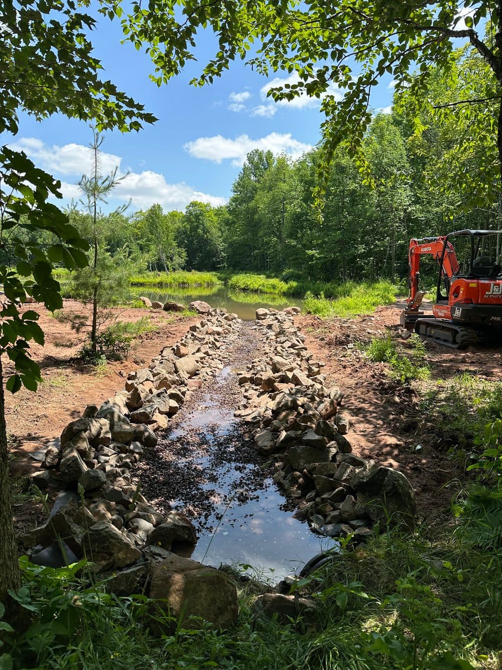 Excavator by a newly constructed rocky stream surrounded by lush greenery and trees.