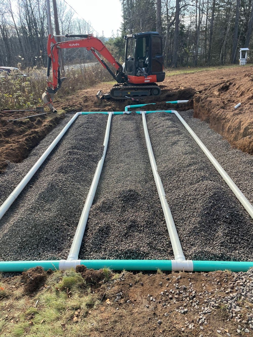 Excavation site with gravel and pipes for drainage system installation. Mini-excavator in background.