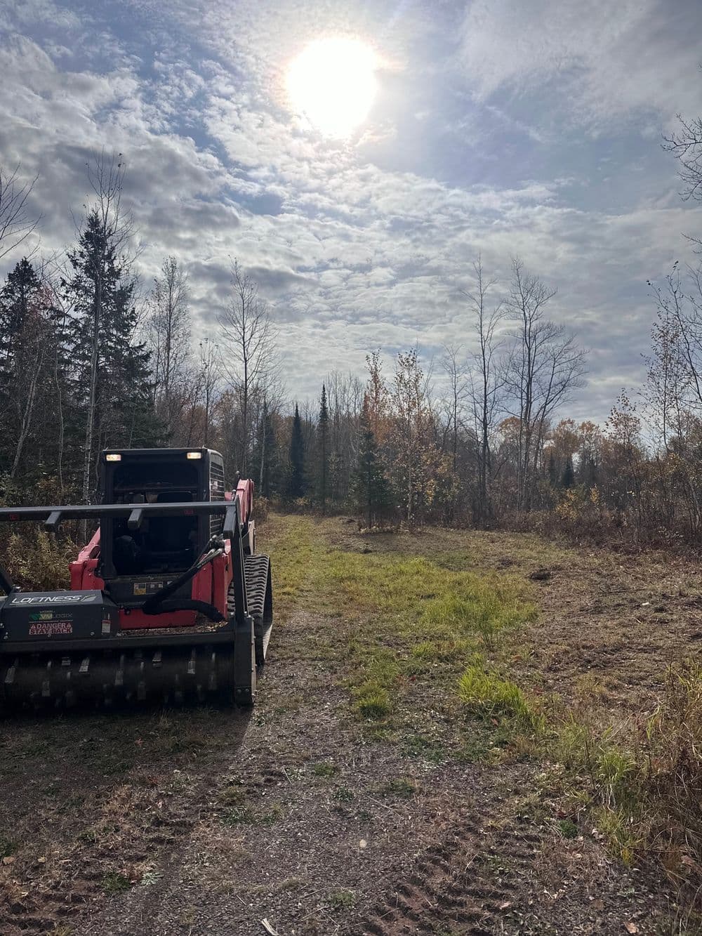 Heavy machinery on a forested path under a bright sun and cloudy sky.