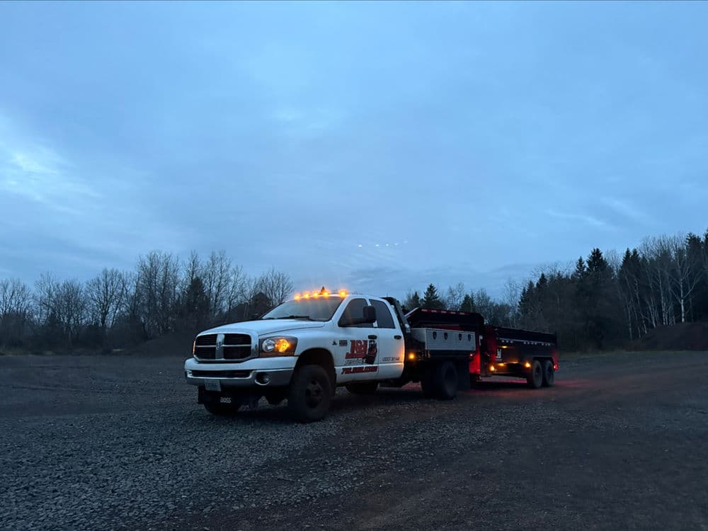 White truck with trailer at dusk, equipped with amber lights, parked on gravel terrain.