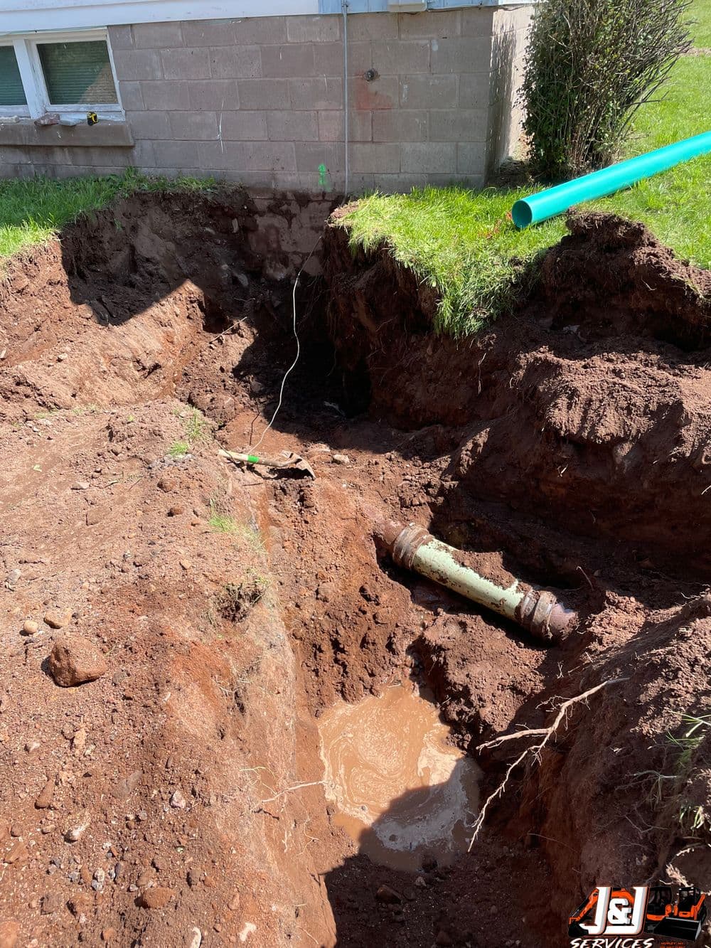 Excavated area revealing a broken pipe surrounded by soil and water near a building.