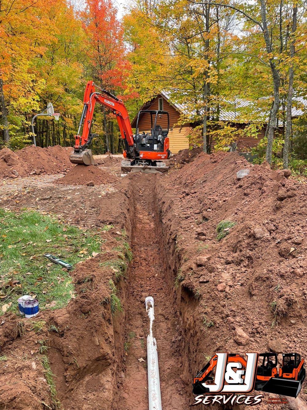 Excavator digging a trench in a wooded area during fall, with a cabin in the background.