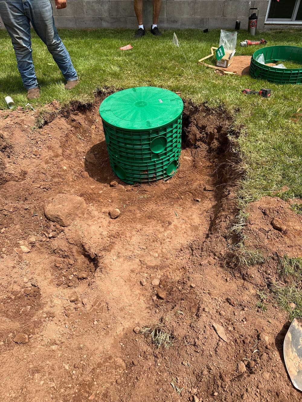 Green drainage basket placed in a dug-out hole in a yard with workers in background.