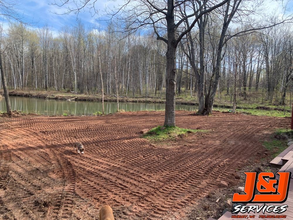 Cleared land near a pond, featuring tire marks in soil and trees in the background.