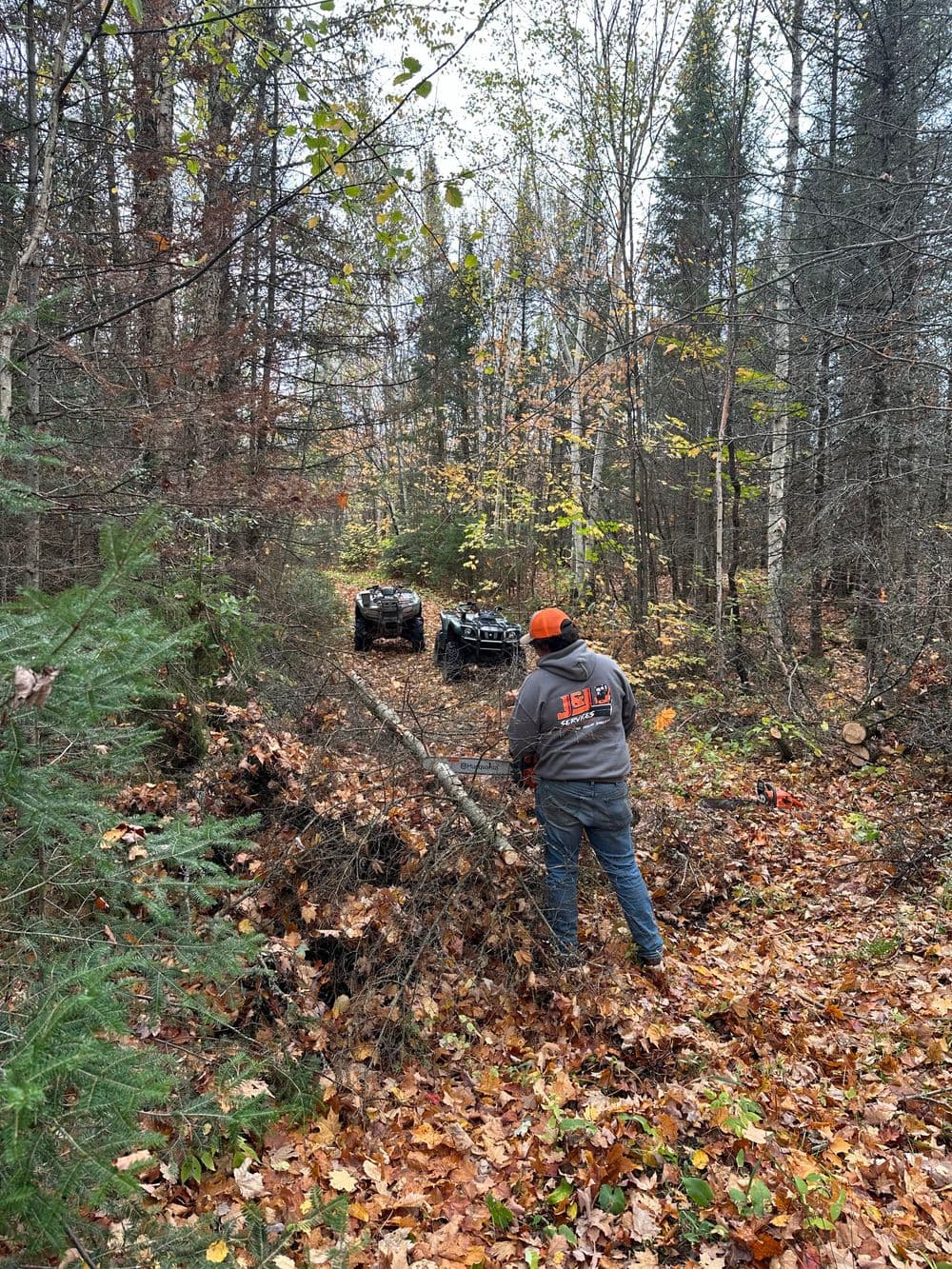 Person clearing fallen branches on a forested trail with two ATVs in the background.
